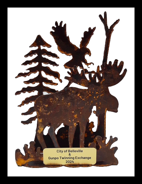 A three dimensional tableau of moose, soaring eagle, and bleak pines in warm, weathered steel. At the base, a plaque in contrasting metal reads "City of Belleville & Gunpo Twinning Exchange 2024" in sans-serif typeface.