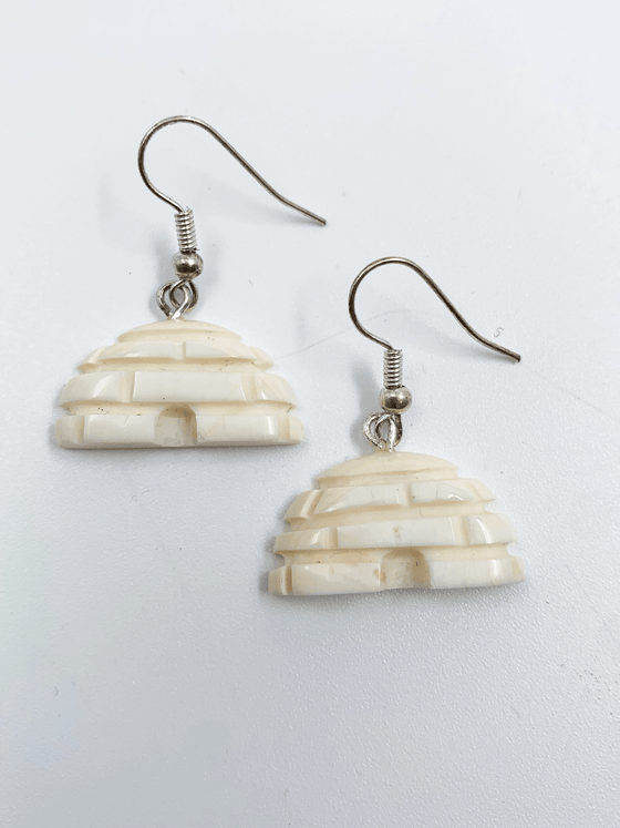 A small pair of off-white igloo earrings. Doors and snow blocks are indented and create texture. Silver earring hooks are attached to the top of the igloos.