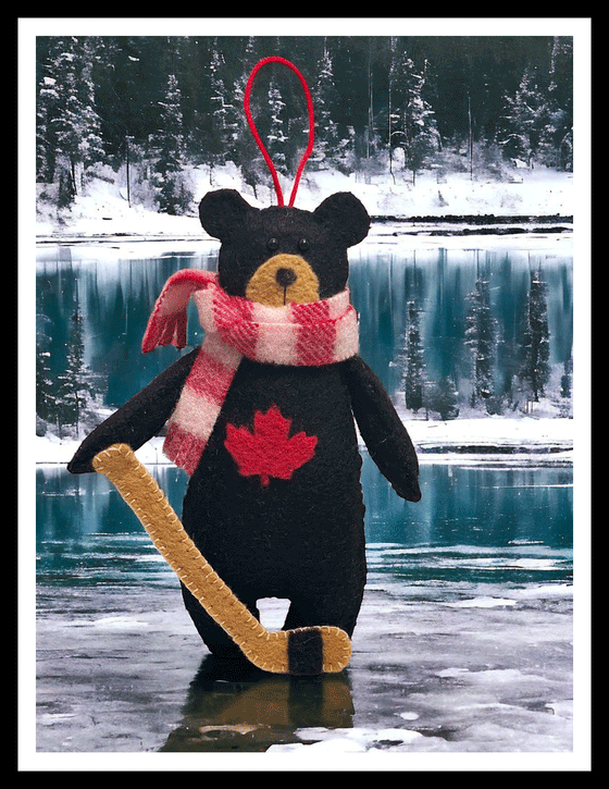 A handmade felted black bear wearing a red and white felt scarf and holding a hockey stick. On its chest, a Canadian red maple leaf is emblazoned.