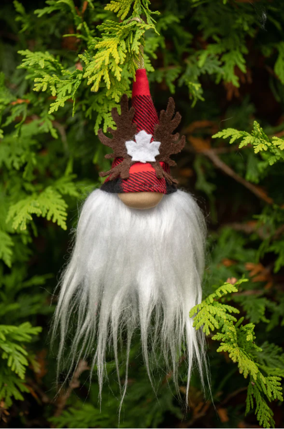 Hanging in a spruce tree, a small uniquely designed character represents a gnome. The gnome figure has a white beard and a red plaid coned hat. The gnomes bulging nose peeks from the plaid hat and is exposed. On the hat there are brown decorative moose antlers with a white maple leaf to match the beard. The ornament hangs from the tip of the hat. Adorable and handcrafted from beautiful fabrics. Home décor with a personality that can be used for the Christmas season or all year round.
