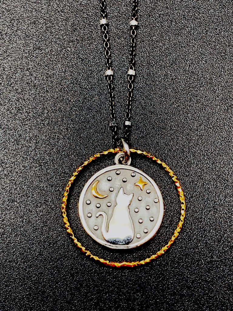 Black chain with thin hexagonal silver rings that are evenly distributed throughout the chain. Pendent is a gold ring that has triangle ridges. A smaller circle with a silver outline and a grey background is inside the ring. There is a cat sitting and facing away in the center of the smaller circle. The cat is surrounded by small silver dots and a gold crescent moon and 4-point star.  