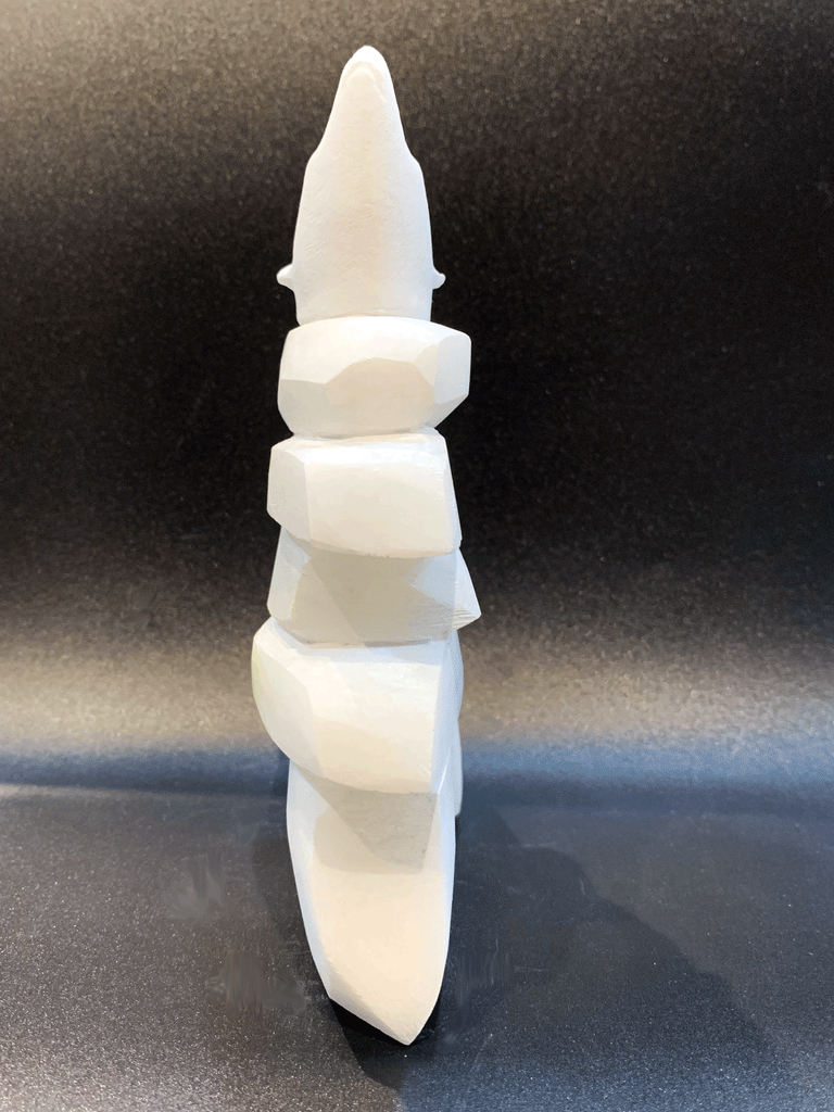 An inukshuk, the top stone of which is a polar bear head looking up, carved in one piece from white alabaster. The piece faces straight ahead in this image. The artist has used a special technique to etch fur texture into the bear and carving marks into the stone of the inukshuk for added realism.