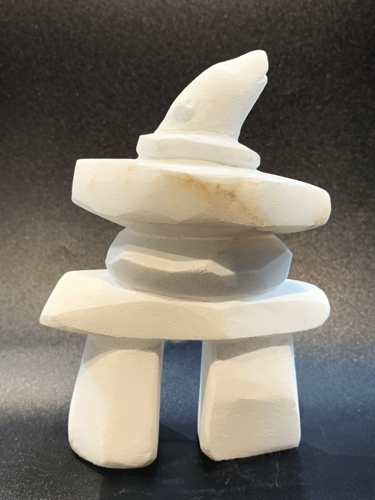 An inukshuk, the top stone of which is a polar bear head looking up, carved in one piece from white alabaster. The piece faces right in this image. The artist has used a special technique to etch fur texture into the bear and carving marks into the stone of the inukshuk for added realism.
