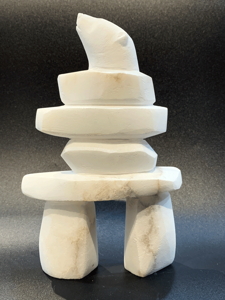 An inukshuk, the top stone of which is a polar bear head looking up, carved in one piece from white alabaster. The piece faces left in this image. The artist has used a special technique to etch fur texture into the bear and carving marks into the stone of the inukshuk for added realism.