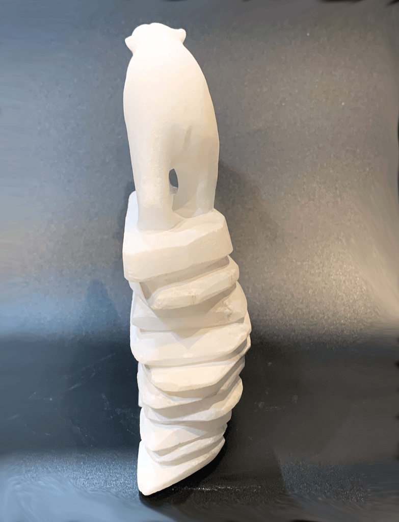 A polar bear standing on top of an inukshuk base, carved in one piece from white alabaster. The polar bear faces away with one forepaw raised. The artist has used a special technique to etch fur texture into the bear and carving marks into the stone of the inukshuk for added realism.
