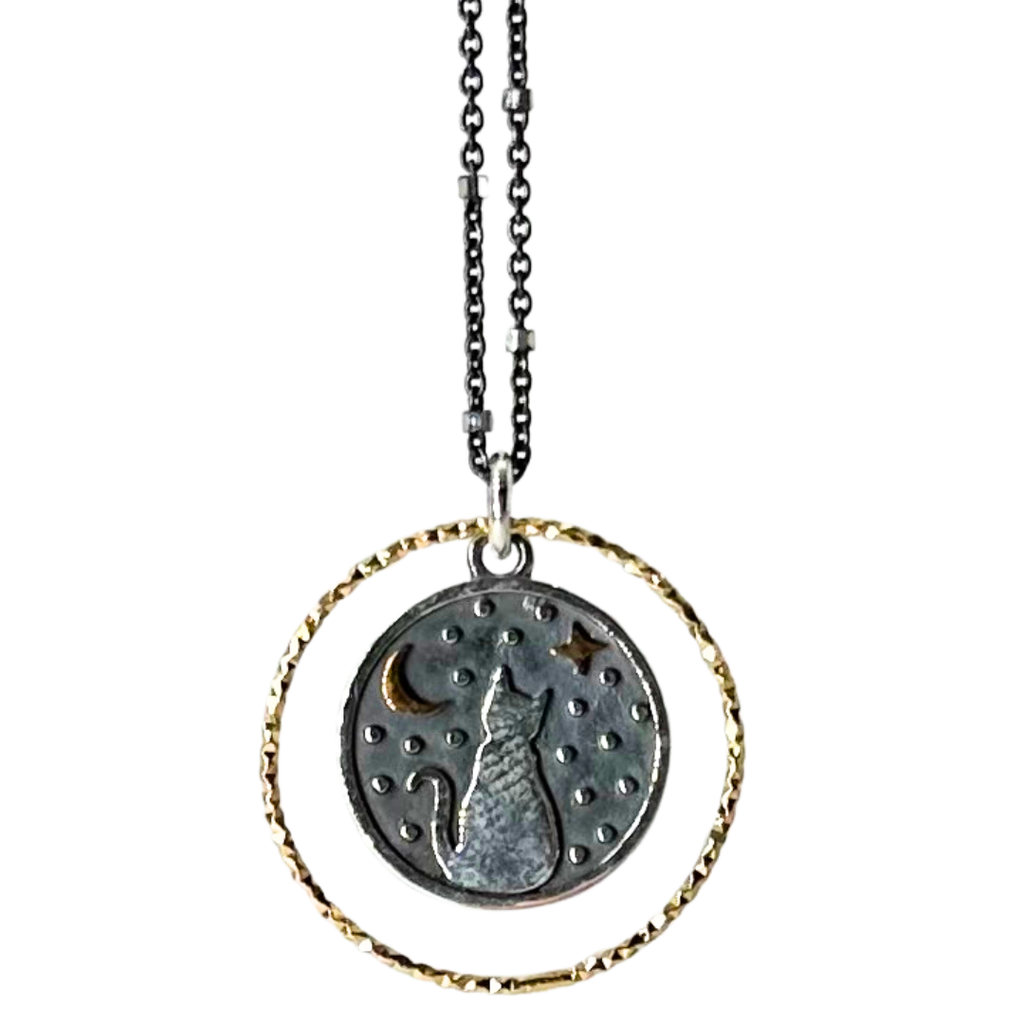 Black chain with thin hexagonal silver rings that are evenly distributed throughout the chain. Pendent is a gold ring that has triangle ridges. A smaller circle with a silver outline and a grey background is inside the ring. There is a cat sitting and facing away in the center of the smaller circle. The cat is surrounded by small silver dots and a gold crescent moon and 4-point star.