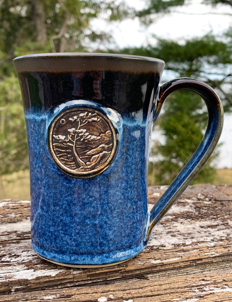 15 oz hand-thrown pottery mug with a vivid black and blue glaze and an embossed windswept pine medallion in the center.
