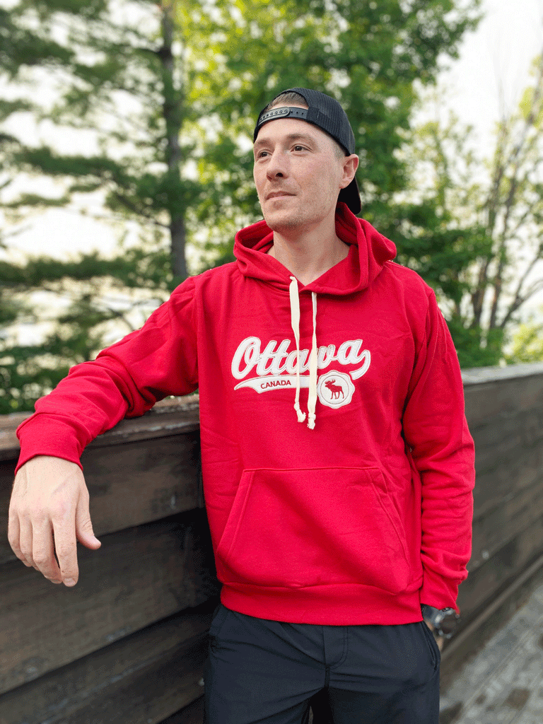 A red hoodie with white drawstrings. Ottawa is written across the chest in a white cursive font, with Canada written in red on a white strip below, next to a red moose on a white circle.
