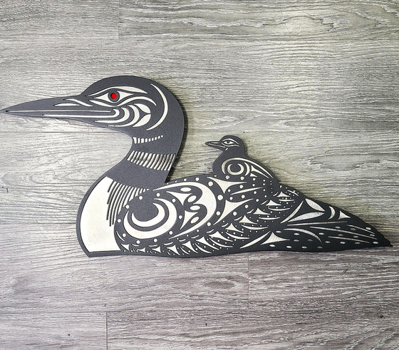 This metal sculpture shows the matte black silhouette of a red-eyed loon with its baby sitting on its back. White-coloured metal shows the details of the loons' beaks, collars, and spotted feathers.