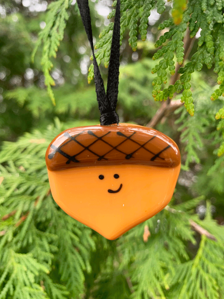 A cute acorn ornament that is made in Canada from fused glass. The acorn is light and dark brown and smiles with a cheerful expression.