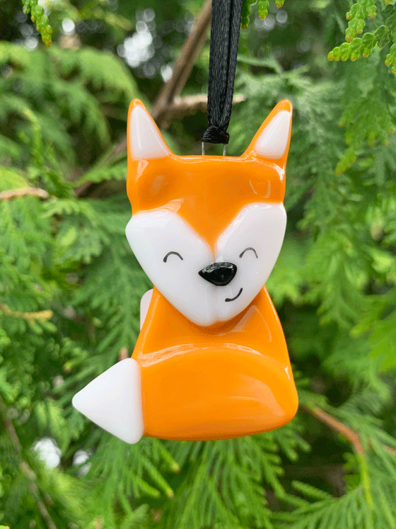 An adorable Canadian made fox ornament, made from fused glass. This piece is orange and white with black accents. The fox has it's tail wrapped around itself and displays a cheerful expression.