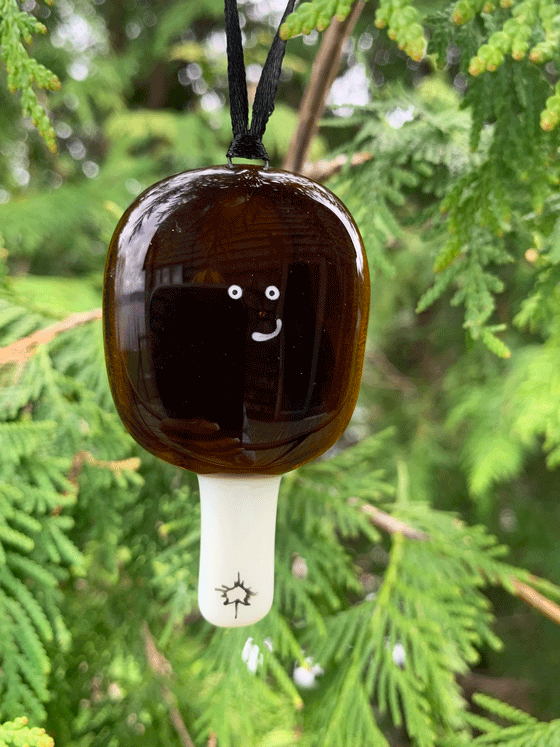 An adorably quirky maple taffy ornament that is Canadian made and made of fused glass. The ornament is dark in colour and round, and the taffy has a cute smiling expression.