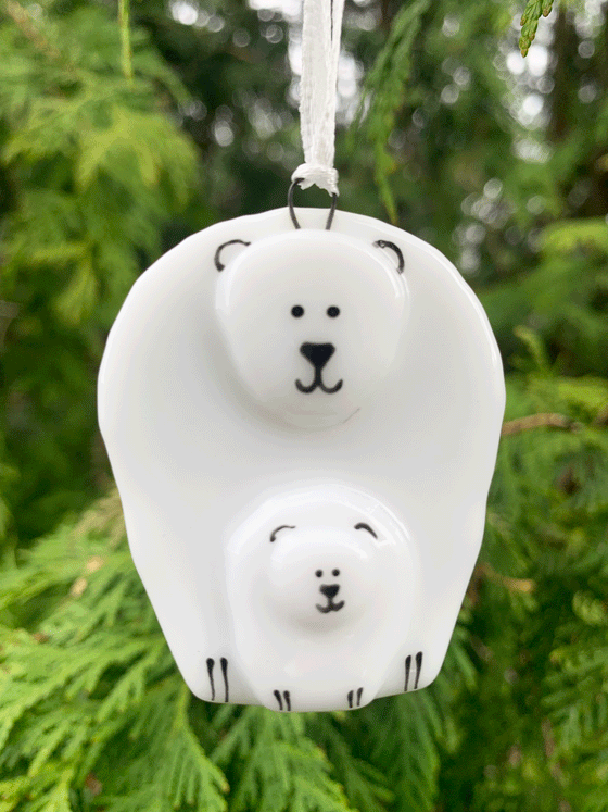 An adorable polar bear ornament that is Canadian made from fused glass. The product is white with black accents and depicts a grown polar bear standing around a baby polar bear. Both bears are smiling serenely.