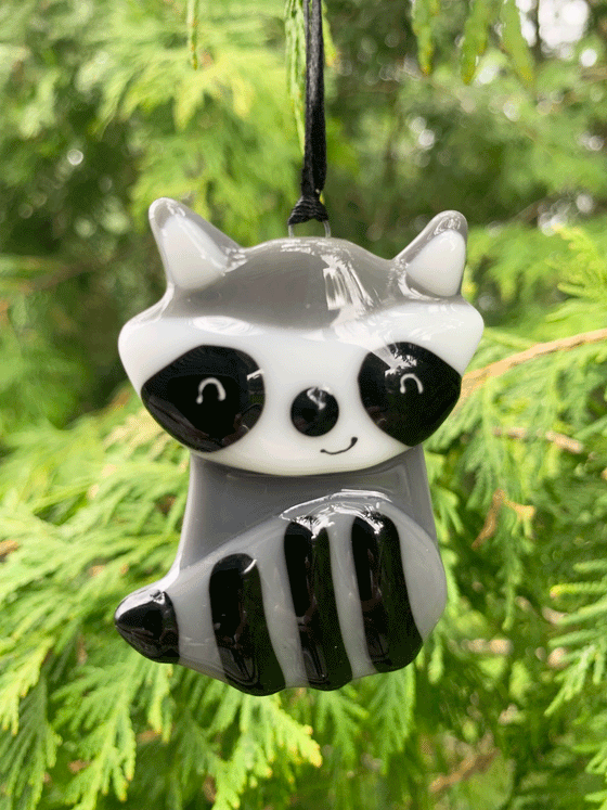 An adorable Canadian made raccoon ornament, made from fused glass. The raccoon is grey, white and black, and it displays a cheerful smile.