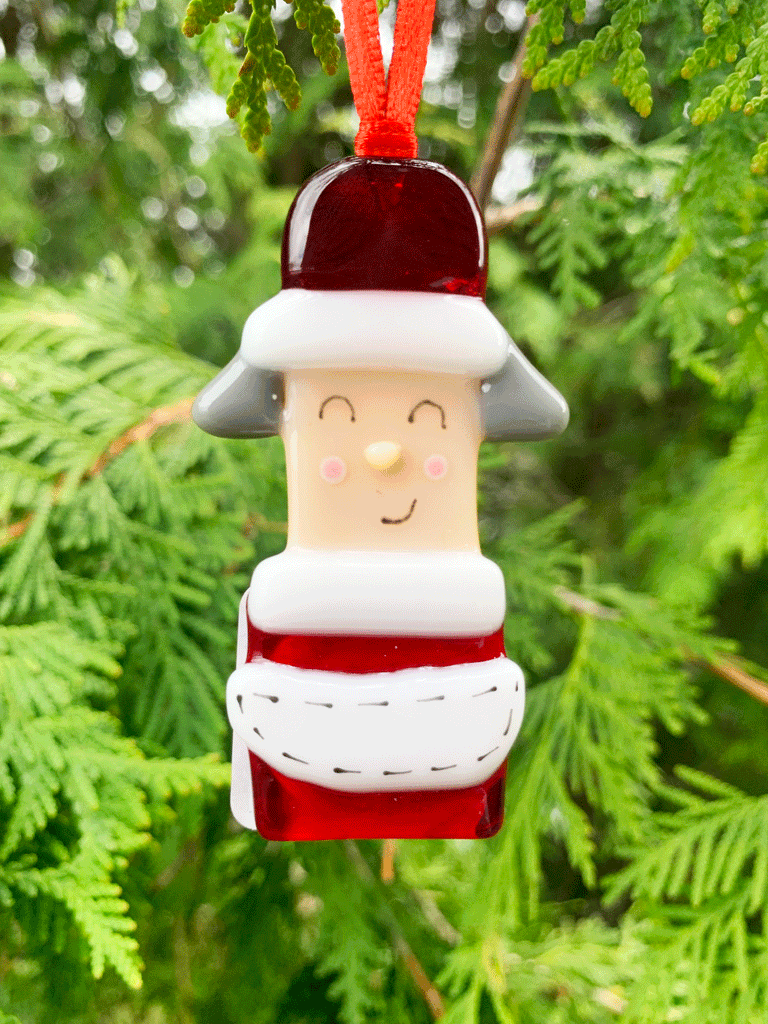 A Mrs. Claus Christmas ornament, made from fused glass, and is Canadian made. Mrs. Claus wears red and white with a matching hat and is kindly smiling.