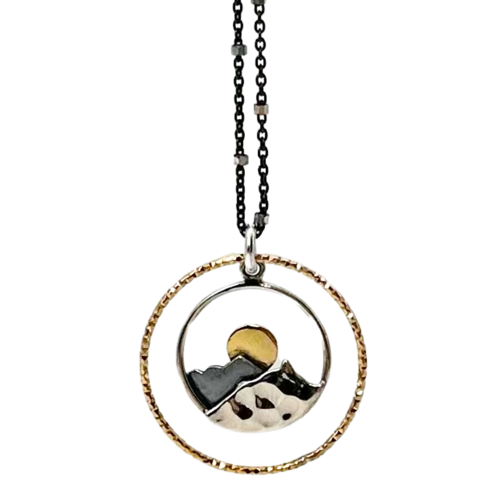 Black chain with thin hexagonal silver rings that are evenly distributed throughout the chain. Pendant is a silver outline of a circle with a bigger gold circle around that. At the bottom of the outline is a silver mountain attached to the outline. The mountain has grooves to make it realistic. Behind the silver mountain is a small, flat, grey mountain with a yellow sun showing behind the peak of the mountain.