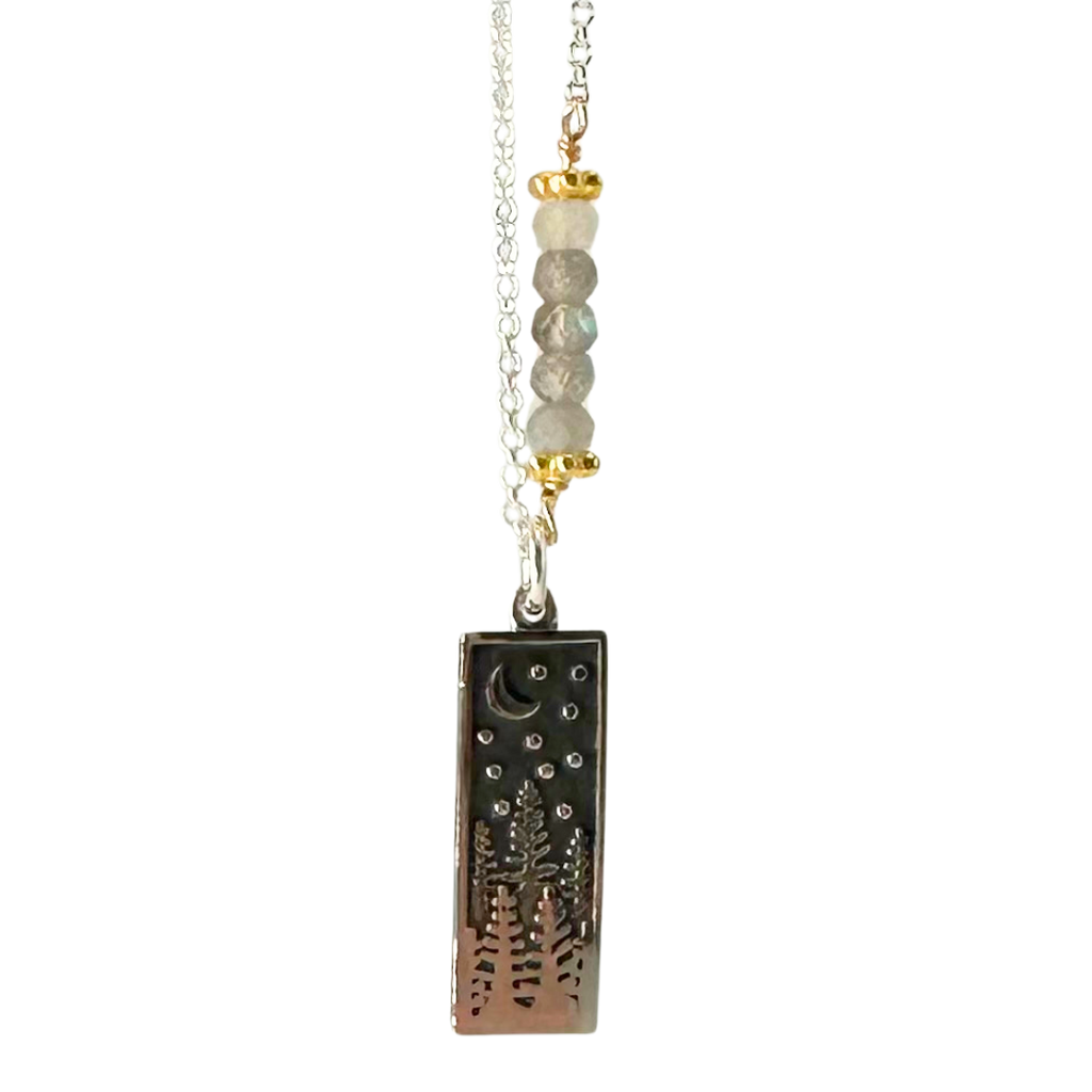 A rectangular silver pendant with impressions of trees, stars, and the moon, hangs on a silver chain with four crystal-like circular beads.