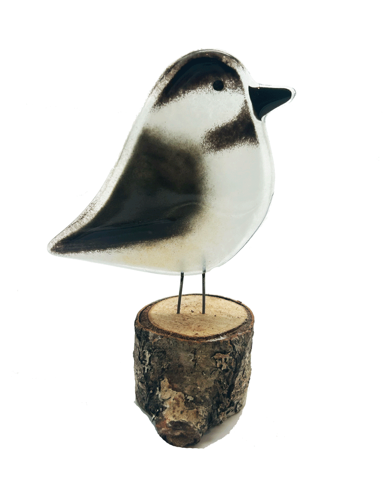 2-D glass black and white Chickadee. Has 2 small metal rods coming from the bottom of it that look like legs. The metal rods are attached to a piece of a tree that looks like a small tree stump. The background is white.