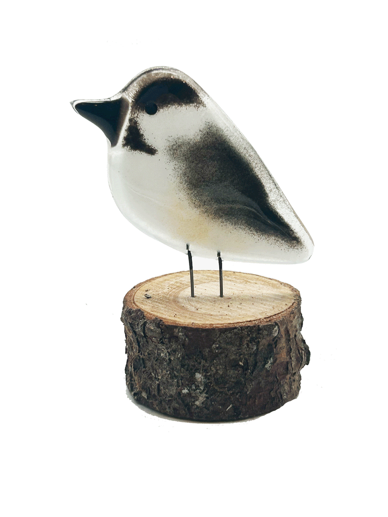 2-D black and white Chickadee. Has 2 small metal rods coming from the bottom of it that look like legs. The metal rods are attached to a piece of a tree that looks like a small tree stump. The background is white.