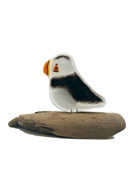 2-D black and white Puffin. Has orange beak and eye. Has 2 small metal rods coming from the bottom of it that look like legs. The metal rods are attached to a piece of a tree that looks like a small tree stump. The background is white.