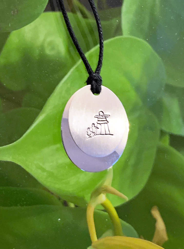 A necklace made of two layers of brushed and polished silver mounted on a leather cord. The base is an oval of bright polished silver. Over that is a smaller circle of brushed silver with a large inukshuk and two smaller human figures etched into the centre.