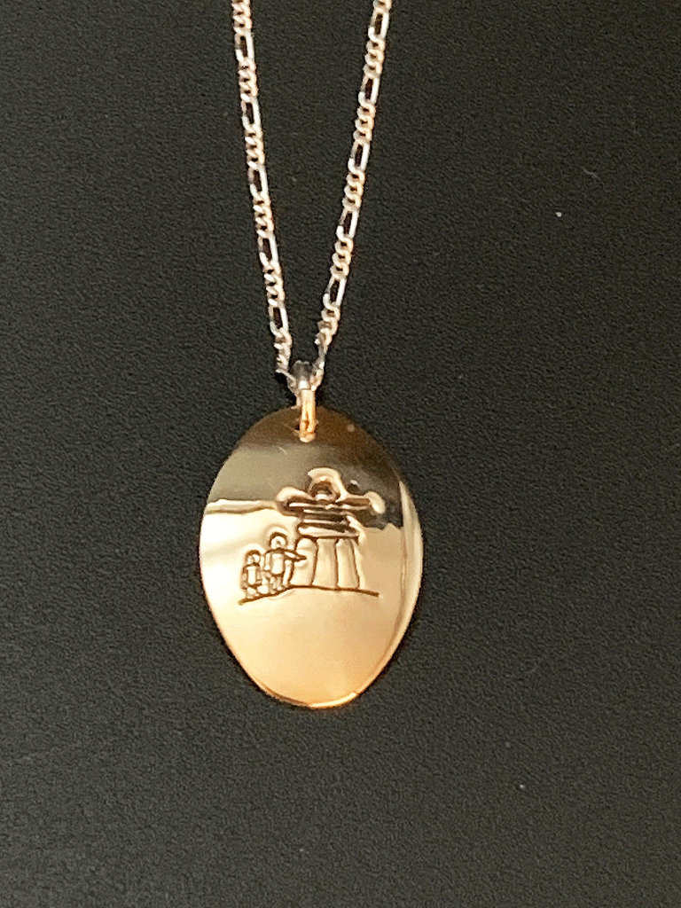 A necklace made of 14 karat gold fill and sterling silver on a gold chain. The base is an oval of bright gold with a large inukshuk with two smaller human figures etched into the centre.