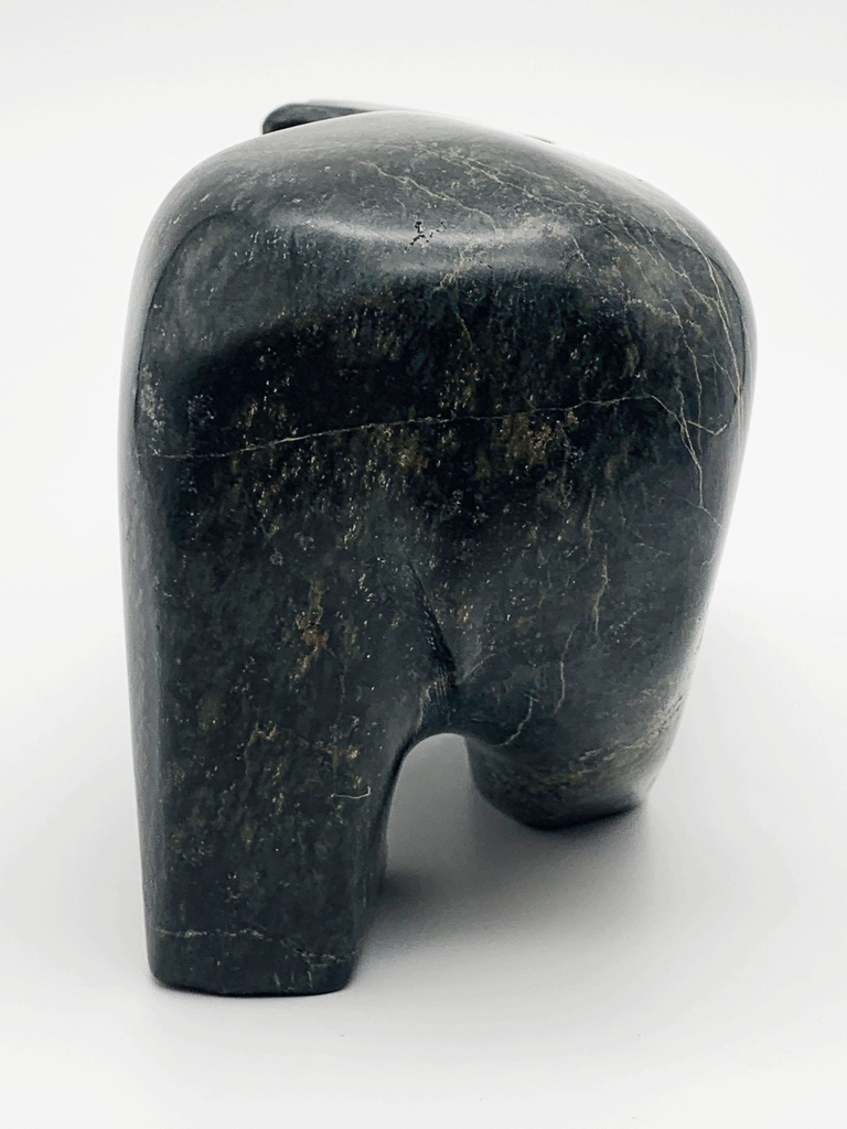 A smiling walking bear carved from very dark green soapstone stands on all fours. Its head is cocked playfully to one side. This bear faces away.