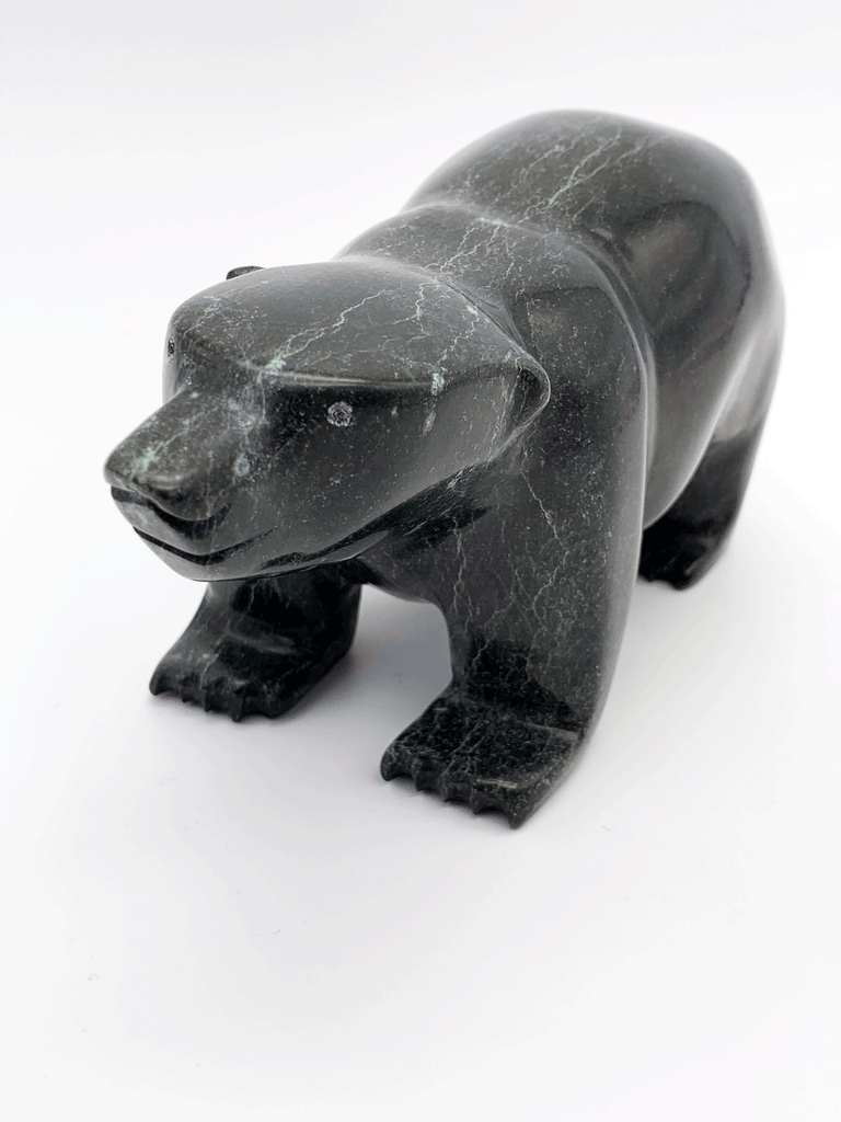 A smiling bear carved from very dark green soapstone stands on all fours, staring straight ahead. This photo is taken at a angle, highlighting the sweet face and sense of scale of the piece.