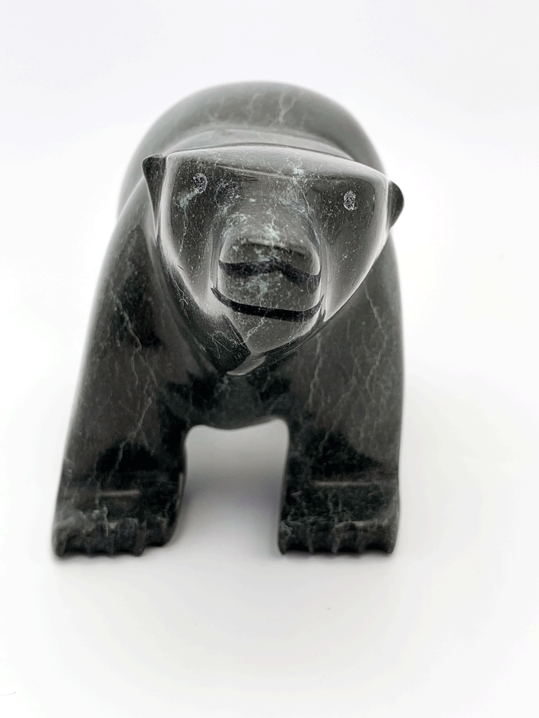 A smiling bear carved from very dark green soapstone stands on all fours, staring straight ahead. This bear faces the viewer.