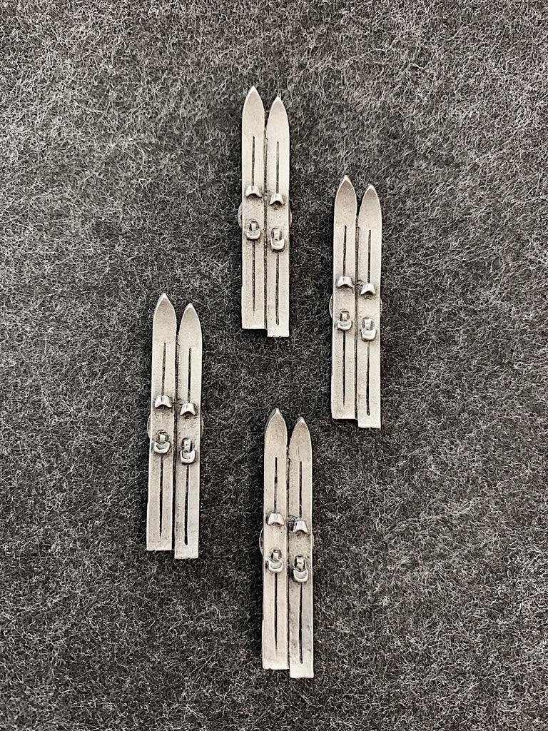 Four pewter magnets in the shape of pairs of skis sitting side by side.