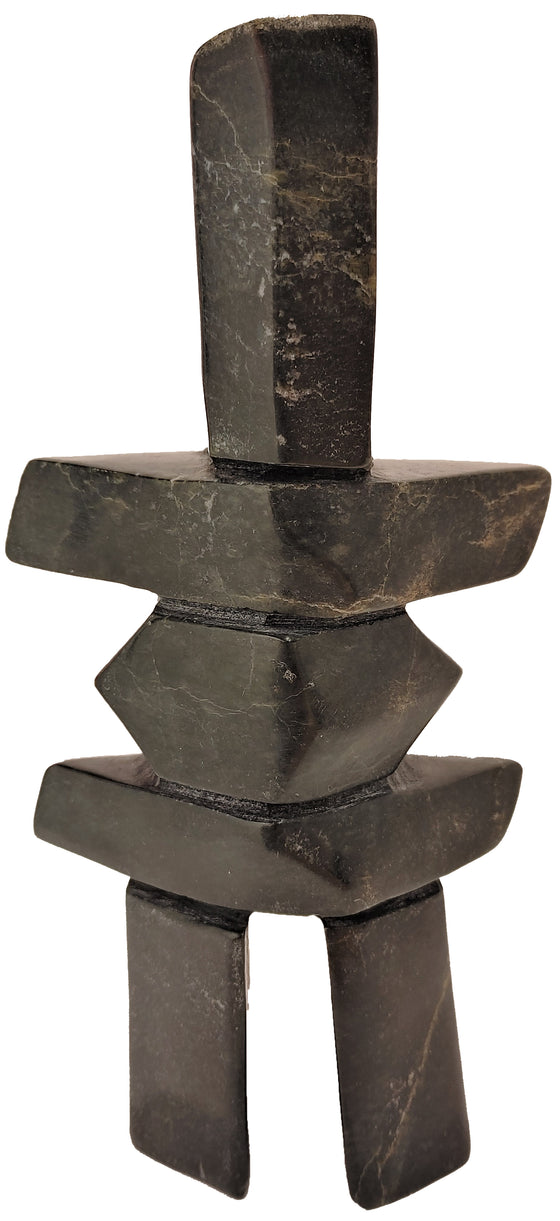 An inukshuk carved from black and brown stone. The legs and head are very tall, while the body is composed of wide slabs forming shoulders and hips, and a rounded stone in the middle.