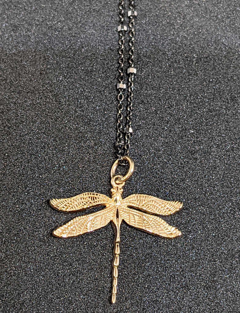 Black chain with thin hexagonal silver rings that are evenly distributed throughout the chain. The pendant is a gold dragonfly with scale detailing on the wings. Necklace is on a black background