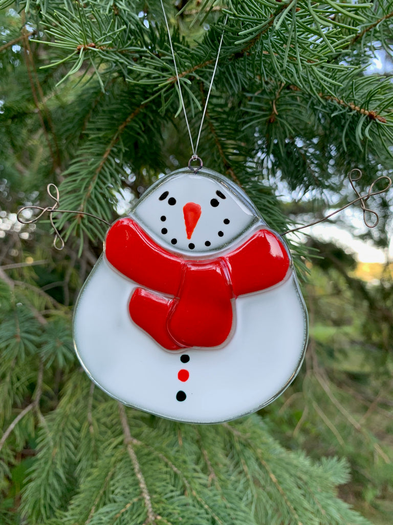 This fused glass ornament depicts a squat, round snowman with a carrot nose and a “coal smile”. He wears a thick red scarf. His arms are made of wire and are outstretched as though asking for a hug.