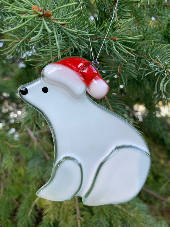This fused glass ornament depicts a polar bear wearing a red and white holiday hat.