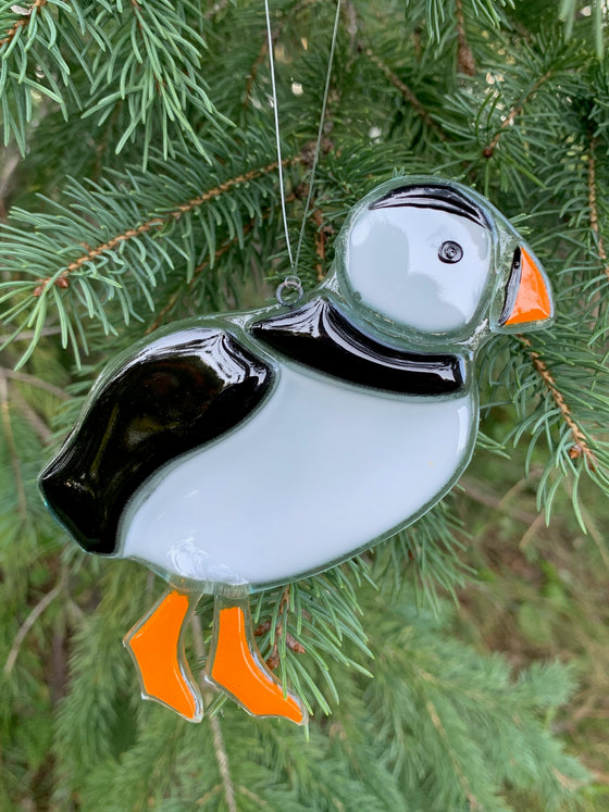 A fused glass ornament showing a puffin. It is black and white with an orange beak and feet.