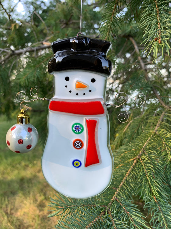 A fused glass ornament depicting a snowman with a carrot nose and a “coal smile”. It is wearing a top hat and a red scarf. Its arms are made of wire and one arm is holding a round ornament.