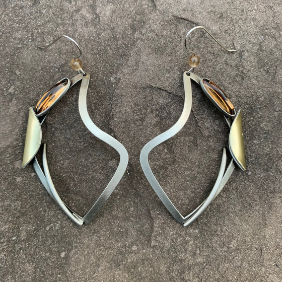 A pair of hook earrings featuring an abstract silver shield shape. Along the outer edge of the shield are a brushed gold semicircle and an oval of amber glass.