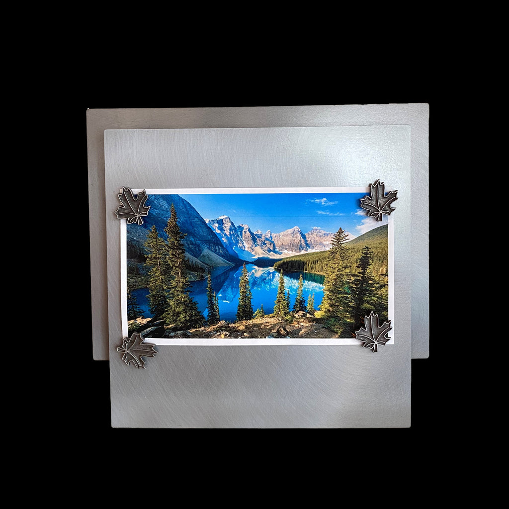 This frame consists of a square, with a rectangle offset upwards and 1/4" behind the square. A photo of a lake with mountains lining it on the left, and pine forest on the right is held on to the frame by four maple leaf shaped magnets. 