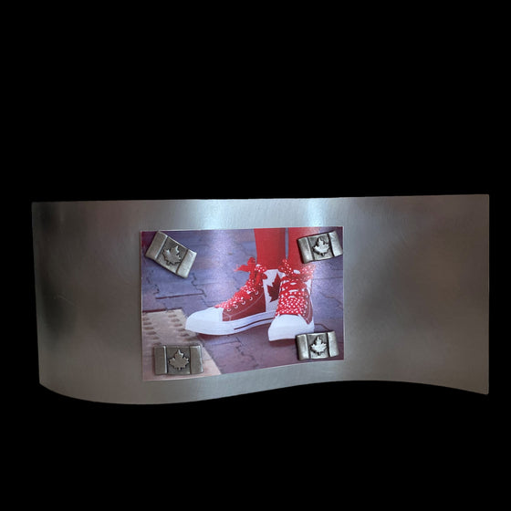 This frame consists of a single rectangular sheet of brushed steel, bent in a wave pattern so the single sheet stands on its own. A photo of someone's Canadian flag shoes is held on by four pewter magnets in the shape of Canadian flags. 