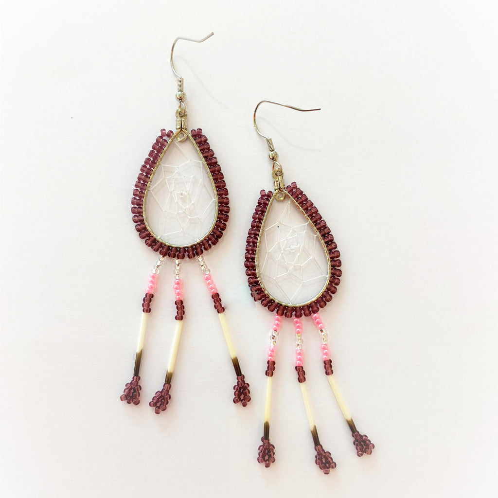 A pair of porcupine quill earrings. The hanging earrings are a tear drop shape hoop with dream catcher style weaving on the inside. Purple beads are woven along the outside of the hoop and hanging off of the bottom are three strands with silver, pink, and purple beads, then a piece of porcupine quill, with more purple beads at the end.