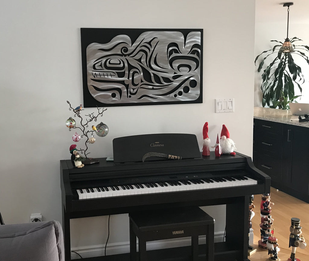 A Coastal Salish Orca wall sculpture hung on a wall. This brushed metal sculpture has been mounted on a black mounting board before being hung to provide contrast between the sculpture and the white wall behind it.