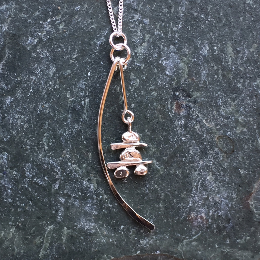 A sterling silver Inukshuk and a long silver crescent hang from a silver chain. The Inukshuk hangs down to the center to the crescent. 