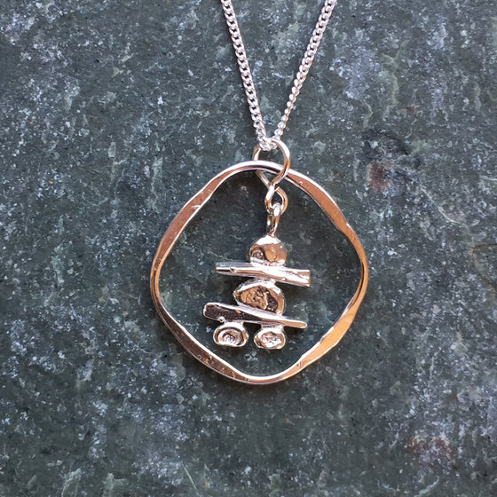 A handmade, sterling silver Inukshuk hanging from a fine silver chain. The Inukshuk is encircled by a beaten silver hoop.