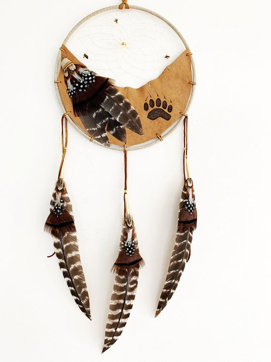 This dream catcher features a crescent of tan leather with a bear’s paw print branded onto it. Above the leather, string is woven in a spiral pattern with small rocks and beads threaded into it. White, black and brown feathers lay across the leather on the left, and hang from three leather strings along the bottom. 