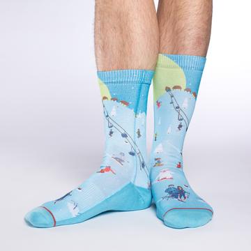 These fun socks feature a cartoon image of a ski slope with people riding a lift, people skiing or snowboarding down the hill, and some shacks at the top of the hill with snow covered pine trees dotted around the slope. The slope is coloured a light blue, while the sky behind is a darker blue with the moon and stars. The sole, toe, and heel of the socks are a darker blue, same as the sky. The active fit socks sport elastic arch bands to contour to your feet and provide support.