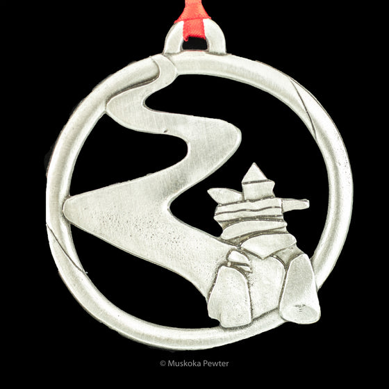 An inukshuk stands at the bottom of a circular frame with a winding path leading beyond the inukshuk tot he top left of the circle. A loop and red ribbon come off the top of the circle. 