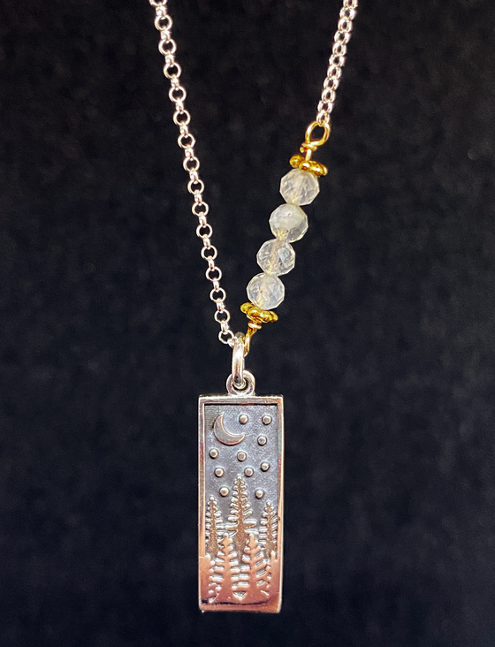 A rectangular silver pendant with impressions of trees, stars, and the moon, hangs on a silver chain with four crystal-like circular beads. 