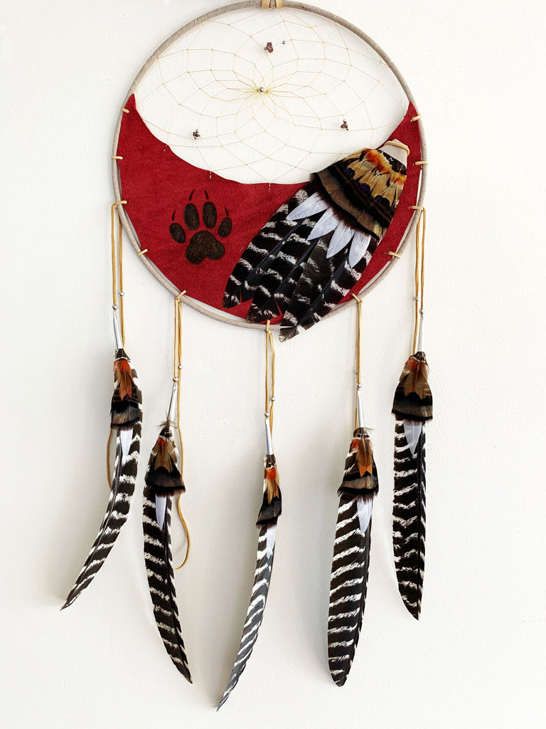 This dream catcher features a crescent of red leather with a wolf’s paw print branded onto it. Above the leather, string is woven in a spiral pattern with small rocks and beads threaded into it. Brown, and white and black feathers lay across the leather on the right, and hang from five leather strings along the bottom. 