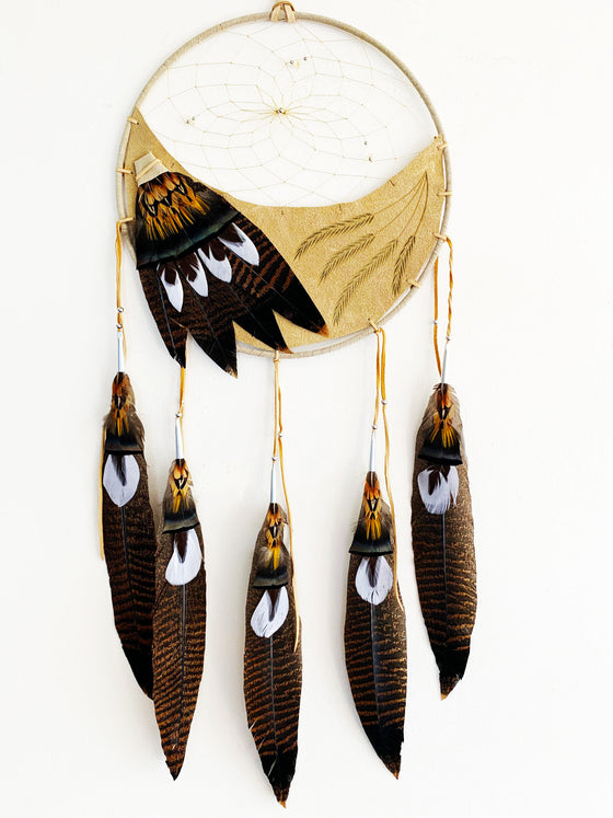 This dream catcher features a crescent of tan leather with grain stalks branded onto it. Above the leather, string is woven in a spiral pattern with small rocks and beads threaded into it. White, and brown and black speckled feathers lay across the leather on the left, and hang from five leather strings along the bottom. 