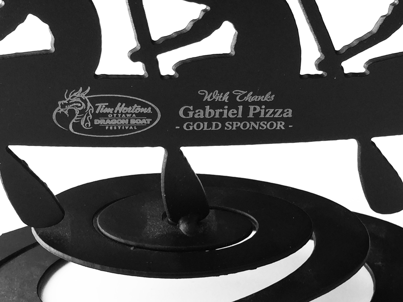A close up of a dragon boat sculpture. The sculpture has been painted matte black. The Tim Hortons Ottawa Dragon Boat Festival logo has been engraved on this piece, as well a thank you to Gabriel pizza for sponsorship. Direct engraving causes the underlying grey metal to show against the black paint.